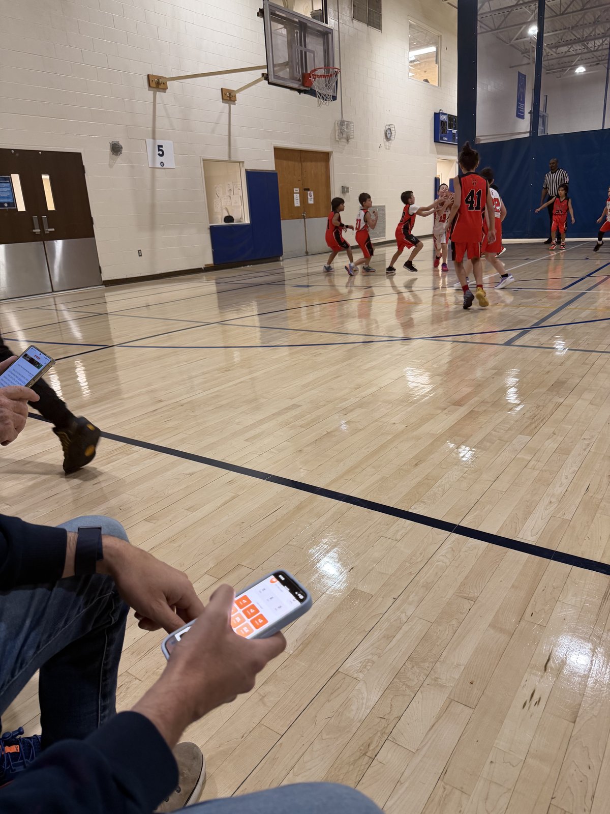 Two parents on the courtside bleachers, each tapping plays into KidGOAT on their phone while their kids' youth basketball game continues on the floor.